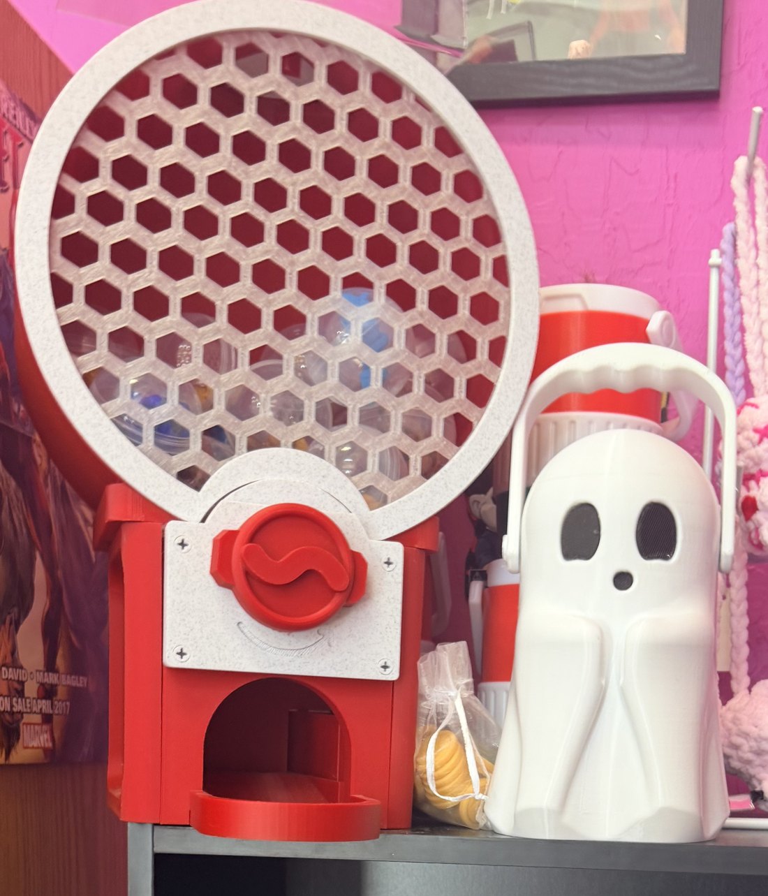 Red 3D printed gumball style vending machine filled with capsules, next to a white 3D printed ghost lantern