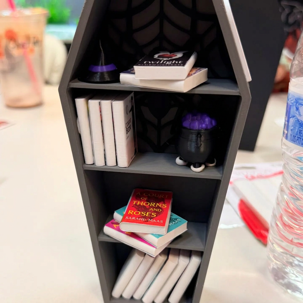 Gray coffin-shaped miniature bookshelf with a black spider, tiny books, and accessories